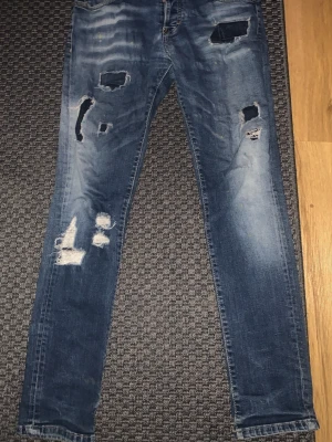 Blå slitna dsquared2 slim fit jeans med hål - Säljer ett par blåa dsquared2 slim fit jeans med flera slitningar och hål framtill. Jeansen har en tvättad look och klassiska bakfickor. Perfekta för en avslappnad och trendig stil. Materialet är denim med stretch för extra komfort.