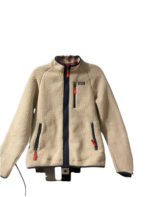 Beige teddyjacka från Patagonia - Mysig beige teddyjacka från Patagonia med marinblå dragkedjor och röda detaljer. Jackan har hög krage, tre fickor med dragkedja och är tillverkad i fluffigt teddy-material som håller dig varm. Perfekt för lager-på-lager och chill höststil. Obs ärmen har gått sönder längst nere vid vänster arm men det är lätt att sy på. 