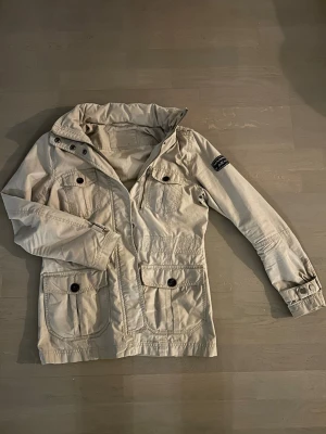 Beige vind jacka från peak performance  - Säljer en beige vindjacka från G-Star RAW med fyra stora fickor framtill, knappar och dragkedja. Jackan har en hög krage och en svart patch på ena ärmen. Perfekt för vår och höst, tillverkad i bomull.
