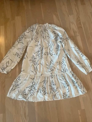 Vit blommig kortklänning med knyt - Söt vit kortklänning med svart blommigt mönster och långa ärmar. Klänningen har volang nertill och knytning vid halsen. Perfekt för vår och sommar, tillverkad i luftig bomull. Aldrig använd!