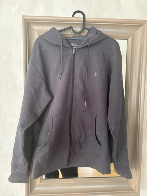 Mörkgrå hoodie från Polo Ralph Lauren - Snygg mörkgrå hoodie med dragkedja från Polo Ralph Lauren. Klassisk design med huva och dragsko, två stora fickor framtill och diskret broderad logga på bröstet. Perfekt för en avslappnad och stilren look.