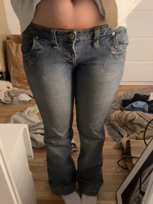 Blå bootcut jeans med dragkedjor - Säljer ett par blå bootcut jeans med slitningar och coola detaljer som dubbla knappar och neonfärgade dragkedjor på bakfickorna. Jeansen har låg midja. Perfekta för dig som gillar en retro vibe! 