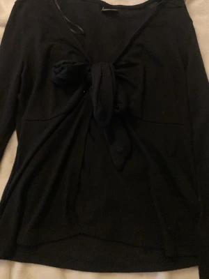 Svart tröja med knyt framtill - Snygg svart tröja med långa ärmar och ett stort knyt framtill som ger en trendig look. Tröjan har en mjuk och tunn stickning, perfekt för lager på lager. Passar till både jeans och kjol för en cool vardagsstil.