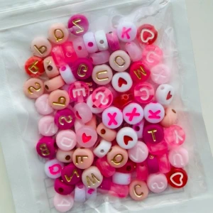 Färgglada bokstavspärlor och symbolpärlor - En påse med mixade pärlor i rosa, vitt och rött med bokstäver, siffror och hjärtsymboler. Pärlorna är runda och cylinderformade, perfekta för att skapa personliga smycken som armband eller halsband. Bokstäver och symboler är tryckta i guld, rosa och rött.