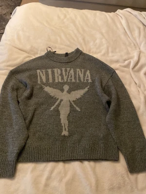 Grå stickad Nirvana-tröja - Stickad grå tröja med Nirvana-tryck och ikonisk ängel på framsidan. Rund halsringning och ribbade muddar vid ärmslut och nederkant. Perfekt för dig som gillar bandmerch och vill ha en cool, avslappnad stil.