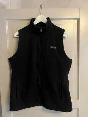 Svart fleeceväst från Patagonia - Klassisk svart fleeceväst från Patagonia med hög krage och dragkedja framtill. Västen har två fickor med dragkedja och en liten logga på bröstet. Perfekt för lager-på-lager och enkel att matcha med olika outfits. Lite liten i storleken! Använt en del