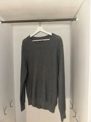 J Lindeberg Grå stickad tröja -STR L  - En stilren grå stickad tröja med rund halsringning och långa ärmar. Tröjan har en enkel, klassisk design med ribbstickade muddar vid ärmslut och nederkant. Perfekt för lager-på-lager och passar till det mesta i garderoben.