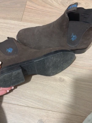 Bruna Chelsea boots från U.S. Polo Assn. - Snygga bruna Chelsea boots i mocka från U.S. Polo Assn. med elastiska paneler på sidorna och blå broderad logga. Klassisk siluett med låg klack och dragflikar både fram och bak. Perfekta för en stilren look.