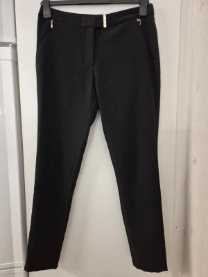 Kostymbyxor - Stilrena svarta kostymbyxor med smal passform och guldfärgade dragkedjor vid fickorna. Byxorna har hög midja och raka ben, perfekta till en dressad look. Materialet ser ut att vara en mjuk och stretchig blandning för extra komfort.