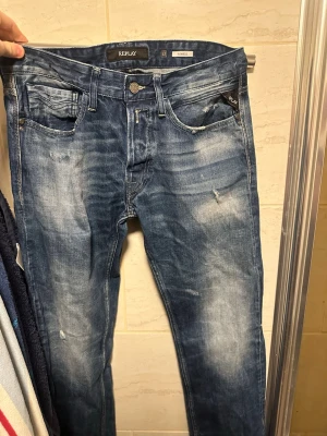 Blå slitna jeans från Replay - Säljer ett par blå jeans från Replay med snyggt slitna detaljer och tvättade partier. Klassisk femficksmodell med knappgylf och smal passform. 