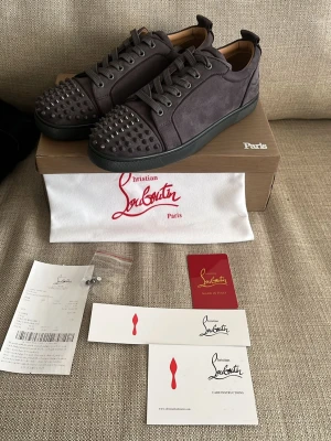Gråa Christian Louboutin sneakers - Säljer ett par gråa sneakers från Christian Louboutin med ikoniska röda sulor och nitar på tån. Skorna är i mocka och har svarta detaljer samt snörning. Perfekta för dig som vill sticka ut med en lyxig och edgy stil.