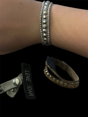 Lene Idar armband med pärlor - Coolt armband från Lene Idar med glittrande pärlor och metallicdetaljer. Armbandet är i grått läder med dubbla rader av pärlor och knäppning med tryckknapp. Perfekt för att ge din outfit lite extra edge. Helt nytt- ny pris 200