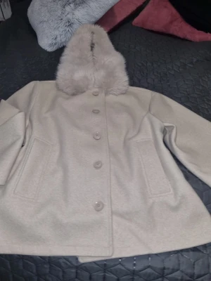 Beige kappa med fuskpäls - Stilren beige kappa med stor fuskpäls på kragen och knäppning framtill. Kappan har två fickor och dekorativa knappar vid ärmsluten. Perfekt för kyliga dagar och ger en lyxig känsla till din outfit. Bara använd ny utan prislapp I storlek L tonåring avdelning 