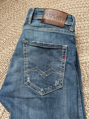Blå jeans från Replay - Säljer ett par klassiska blå jeans från Replay med snygga detaljer på bakfickan och läderpatch i midjan. Jeansen har raka ben och är tillverkade i slitstarkt denim. Perfekt för dig som gillar en tidlös och cool look.