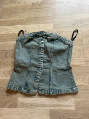 Denim tubtopp med knappar - Säljer en snygg denim tubtopp med svarta axelband och knappar framtill. Toppen har en croppad passform och markerade sömmar som ger en cool look. Perfekt att styla med höga jeans eller kjol för en trendig outfit.