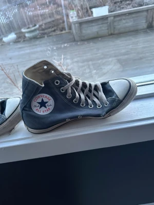 Converse All Star blå high tops - Klassiska Converse All Star high tops i mörkblå canvas med vita gummisulor och tå, samt vita snören. Ikonisk rund logga på sidan och metallöljetter för snörningen. Perfekta för en avslappnad och cool streetstil.