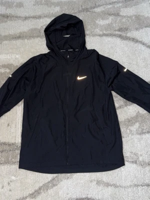 Nike miler  - Svart vindjacka från Nike med huva och hel dragkedja. Jackan har reflekterande Nike-logga på bröstet och ärmen samt två sidofickor. Perfekt för löpning eller utomhusaktiviteter. Lätt och smidig design. Litet hål där bak 