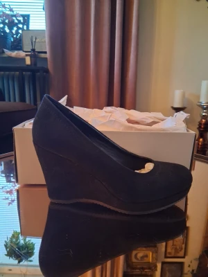 Svarta kilklackade pumps - Snygga svarta pumps med hög kilklack och rundad tå. Skorna har en stilren design och är tillverkade i ett material som ser ut som mocka. Perfekta för dig som vill ha både höjd och stabilitet i din look.