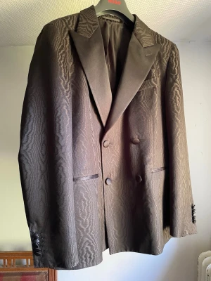 Brun/Svart mönstrad kavaj från Hugo - Snygg brun kavaj från Hugo med diskret vågmönster i tyget. Dubbelknäppt modell med breda slag och klädda knappar både fram och vid ärmslut. Kavajen har två fickor fram och är helfodrad för en lyxig känsla.