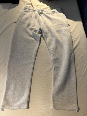 Ljusgrå mjukisbyxor med dragsko - Säljer ett par ljusgrå mjukisbyxor med dragsko i midjan och raka ben. Byxorna har sidofickor och en bakficka. Tillverkade i mjukt sweatshirtmaterial, perfekta för chill dagar hemma eller när du vill vara bekväm.