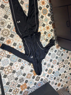 Svart träningsset från Under Armour - Säljer ett svart träningsset från Under Armour med vita detaljer. Setet består av en hoodie med dragkedja och byxor, båda med logga på bröst och ben. Materialet är stretchigt och passar perfekt för träning eller chill. Snygg passform och sportig look.