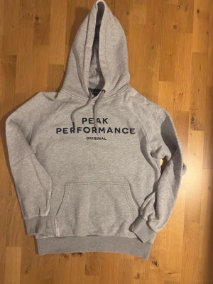 Grå hoodie Peak Performance - Snygg grå hoodie från Peak Performance med svart broderad logga på bröstet och texten 'ORIGINAL' under. Klassisk känguruficka framtill, ribbade muddar och justerbar huva med dragsko. Perfekt för en avslappnad och sportig stil.