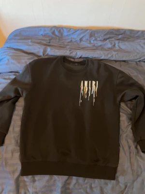 Svart Amiri sweatshirt med färgstänk - Cool svart sweatshirt från Amiri med färgstänk och stor logga både fram och bak. Tröjan har rund halsringning, ribbade muddar och en avslappnad passform. Perfekt för dig som gillar streetwear och vill sticka ut med en unik design.