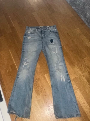Ljusblå slitna bootcut jeans - Säljer ett par ljusblå bootcut jeans med slitningar och hål på både fram- och baksida. Jeansen har klassisk femficksmodell och är tillverkade i denim. Perfekta för en avslappnad och trendig look.