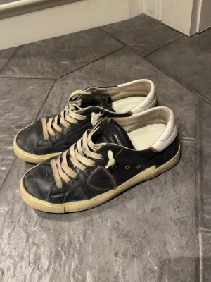 Svarta Philippe Model - Snygga svarta sneakers från Philippe Model med vita detaljer och beige snören. Skorna har en låg silhuett, vit sula och diskret logga på sidan. Materialet är en mix av läder och textil, med kontrasterande vit hälkappa och röda sömmar.