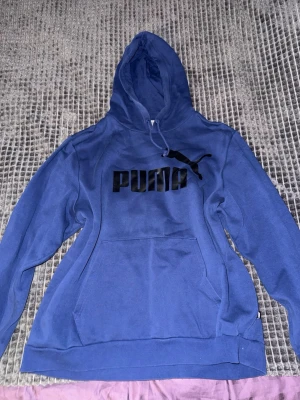 Blå hoodie från Puma med logga - Säljer en blå hoodie från Puma med stor svart logga och text på bröstet. Hoodien har huva med dragsko och en stor magficka framtill. Tillverkad i mjukt bomullsmaterial som är skönt att ha på sig. Perfekt för dig som gillar streetwear och sportig stil.