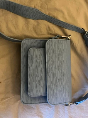 Ljusblå axelväska med struktur - Snygg ljusblå axelväska i konstläder med präglad struktur och justerbar bred axelrem. Väskan har två framfickor med lock och silverfärgade metalldetaljer. Perfekt för dig som vill ha en stilren och modern accessoar.