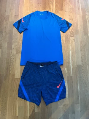 Blått träningsset från Nike - Snyggt blått träningsset från Nike med t-shirt och shorts. T-shirten har svarta och blå mönstrade detaljer på ärmarna samt Nike-logga i orange. Shortsen är blå med svart mönster och har orange Nike-logga. Perfekt för träning eller sport.