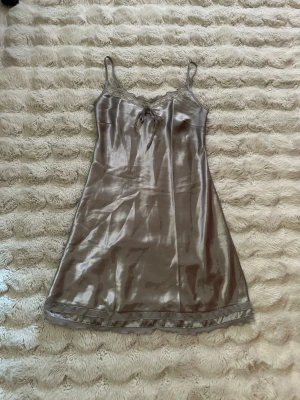 Nattlinne/neglige - Satin Nattlinne i en vacker grå nyans med justerbara axelband. Nattlinnet har fina spetsdetaljer längs urringningen och en söt rosett framtill. Nederkanten är prydd med volanger, vilket ger en charmig touch. Baktill finns en liten öppning med en knappstängning.