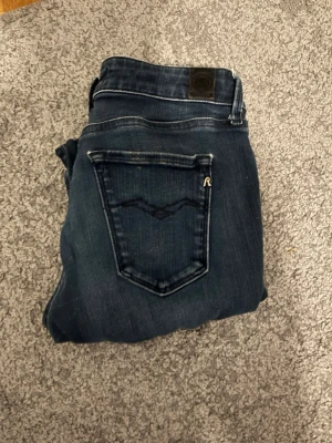Mörkblå skinny jeans från Replay l28 w27 - Snygga mörkblå skinny jeans från Replay med klassisk femficksdesign och diskreta slitningar. Jeansen har en stilren bakficka med broderad detalj och läderpatch i midjan. Perfekta för dig som gillar en smal passform och tidlös look.