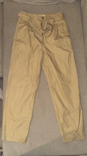 Beiga chinos med bälte - Snygga beiga chinos med tillhörande bälte och hög midja. Byxorna har raka ben, två sidofickor fram och två passpoalfickor bak. Tillverkade i ett mjukt bomullstyg som känns skönt mot huden. Perfekta för en avslappnad och trendig look.