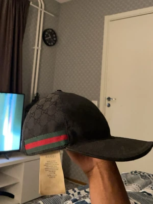 Svart (ÄKTA) gucci keps - Snygg svart keps från Gucci med klassiska GG-monogram över hela kepsen och de ikoniska gröna och röda ränderna på sidan. Kepsen har böjd skärm och är tillverkad i ett slitstarkt tygmaterial. Perfekt för dig som vill ha en exklusiv och trendig accessoar. 