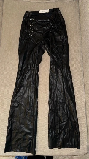 Svarta lace-up byxor i skinnimitation - Säljer ett par svarta byxor i skinnimitation från Bershka med snörningsdetaljer framtill. Byxorna har utsvängda ben och hög midja, vilket ger en edgy och trendig look. Perfekta för dig som vill sticka ut med en cool stil.