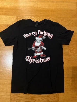 Svart jul-tshirt med tryck - Svart t-shirt med korta ärmar och tryck av en julig tomte med texten 'Merry f*cking Christmas' i vitt och rött. Perfekt för dig som vill sticka ut på julfesten. Skön passform och klassisk rund hals.
