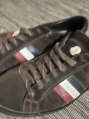 Bruna Moncler sneakers med ränder - Snygga bruna sneakers från Moncler med klassiska röda, vita och blå ränder på sidan samt Moncler-logga på plösen. Skorna har matchande bruna skosnören med röda och vita detaljer. Tillverkade i läder med gummisula. Inte bra alls men kan reparerats enkelt. 