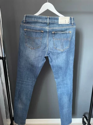 Blå jeans från Tiger or sweden - Snygga blå jeans från Tiger of sweden med klassisk femficksdesign och raka ben. Jeansen har ljus tvätt och kontrastsömmar, samt en vit patch med logga bak i midjan. Perfekta för en avslappnad och trendig look.