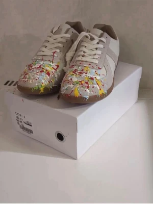Maison margielas med färgstänk  - Unika vita sneakers med beige detaljer och färgstänk i rött, gult och grönt över tån. Skorna har klassisk låg siluett, vita snören och brun sula. Materialet är en mix av läder och mocka för en cool och trendig look.