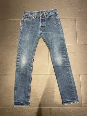 Blå raka jeans från Levi's - Klassiska blå jeans från Levi's med raka ben och fem fickor. Jeansen har en lätt tvättad look med slitningar på knän och benslut. Tillverkade i bomullsdenim och har den ikoniska läderpatchen bak i midjan. Trasig söm på ena bakfickan som manninte tänker på, ställ frågor vid funderingar!