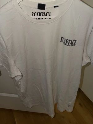 Vit Scarface t-shirt med tryck - Vit t-shirt med Scarface-tryck på bröstet och stort motiv på ryggen. Klassisk rund hals och korta ärmar. Materialet är mjuk bomull och designen har en cool grafisk stil med filmtema.