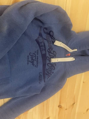Blå Superdry hoodie med tryck - Säljer en blå hoodie från Superdry med stort tryck på bröstet och japanska tecken. Hoodien har vit dragsko i huvan och en klassisk känguruficka framtill. Perfekt för dig som gillar streetwear och vill ha något bekvämt och snyggt.
