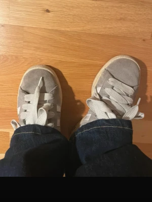 Adidas campus - Snygga grå sneakers från Adidas i mocka med klassiska vita ränder på sidorna och vit sula. Skorna har breda vita skosnören och låg profil, perfekt för en avslappnad streetstil.