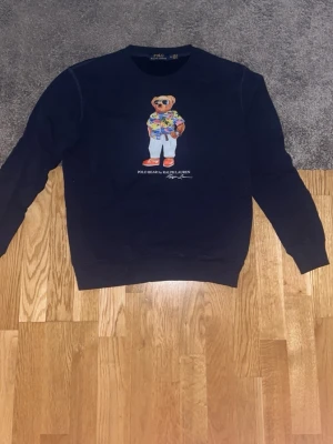 Mörkblå sweatshirt Polo Ralph Lauren - Mörkblå sweatshirt från Polo Ralph Lauren med ikoniska Polo Bear-trycket på bröstet. Rund halsringning och ribbade muddar vid ärmslut och nederkant. Perfekt för dig som gillar streetwear och klassisk stil. Storlek S och M