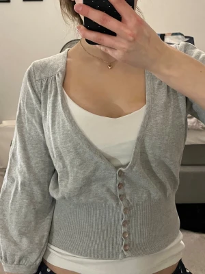 Ljusgrå kofta med knappar - Snygg ljusgrå kofta med djup V-ringning och dekorativa knappar framtill.  (Okänt märke) Modellen har trekvartsärmar och bred ribbad midja. Superskön och inga synliga fläckar eller liknande💕 storlek S men tror även den passar M då materialet är stretchigt 