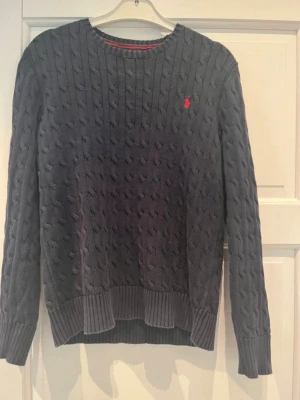 Mörkblå kabelstickad tröja Polo Ralph Lauren - Kabelstickad mörkblå tröja från Polo Ralph Lauren med rund halsringning och röd broderad logga på bröstet. Tröjan har ribbstickade muddar vid ärmslut och nederkant. Perfekt för vintern och den har en klassisk stil.