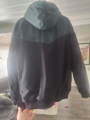 Svart och mörkgrön vindjacka med huva - Snygg vindjacka i svart och mörkgrönt med dragkedja framtill och två fickor med dragkedja. Jackan har en justerbar huva och muddar vid ärmslut och nederkant. Perfekt för blåsiga dagar och enkel att matcha med olika outfits. Finns något hål på ärmen annars bra skick 