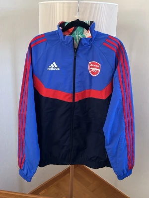 Arsenal Adidas vind/träningsjacka - Arsenal vind/träningsjacka från Adidas storlek M.  Jackan är alltså 2 jackor i 1. Om man vänder jackan ut och in så blir det en annan jacka i en annan färg. Så ena sidan är blå o har röda detaljer och adidas ränder längs ärmarna. Andra sidan är istället mönstrad i röd o grön med vita adidas detaljer o Arsenal märke. Den har hel dragkedja framtill och resår vid ärmslut och midja. Perfekt för all typ av användning. 